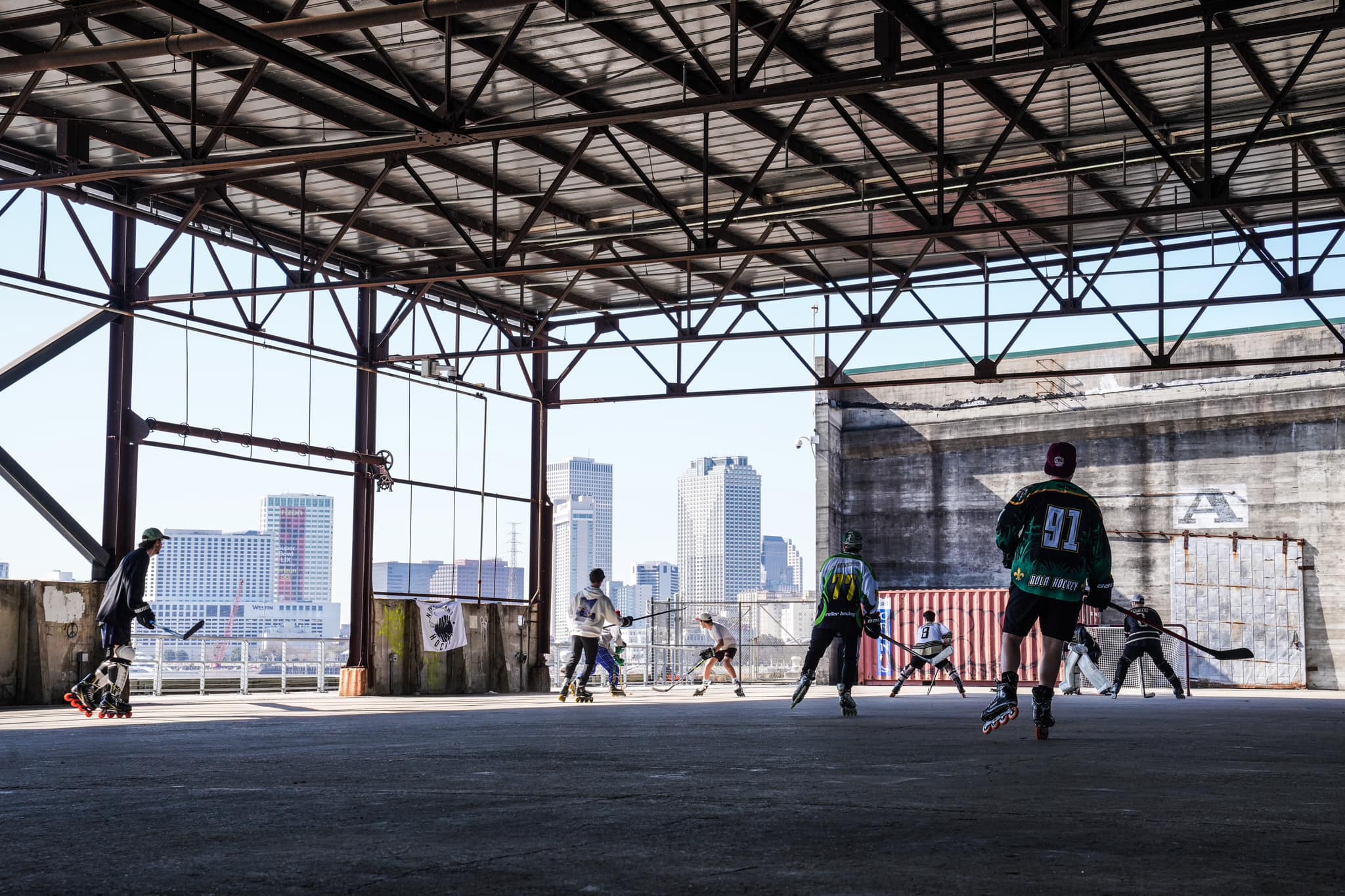 Players skating roller hockey inside the Crescent Park warehouse with the New Orleans skyline visible in the background